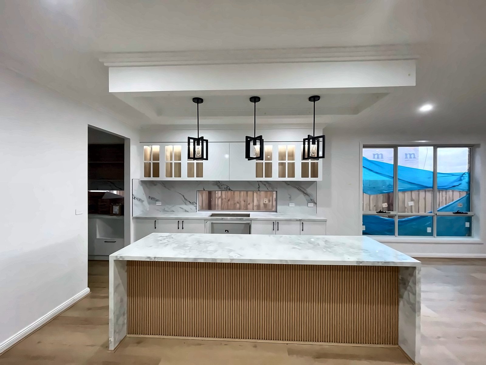 White kitchen with glass-front display overheads, a fluted timber island and marble-look tops, by Maya Cabinets in Melbourne
