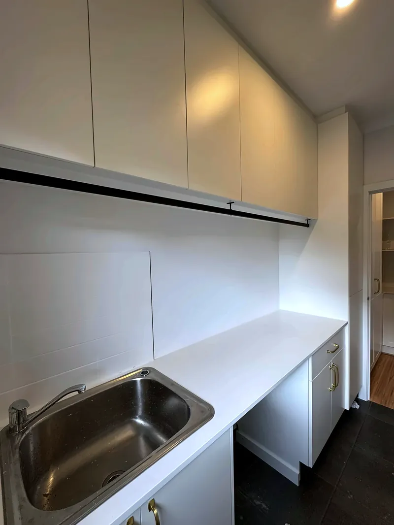 White laundry with a stone bench, tub, overhead cupboards and a hanging rail, by Maya Cabinets in Melbourne